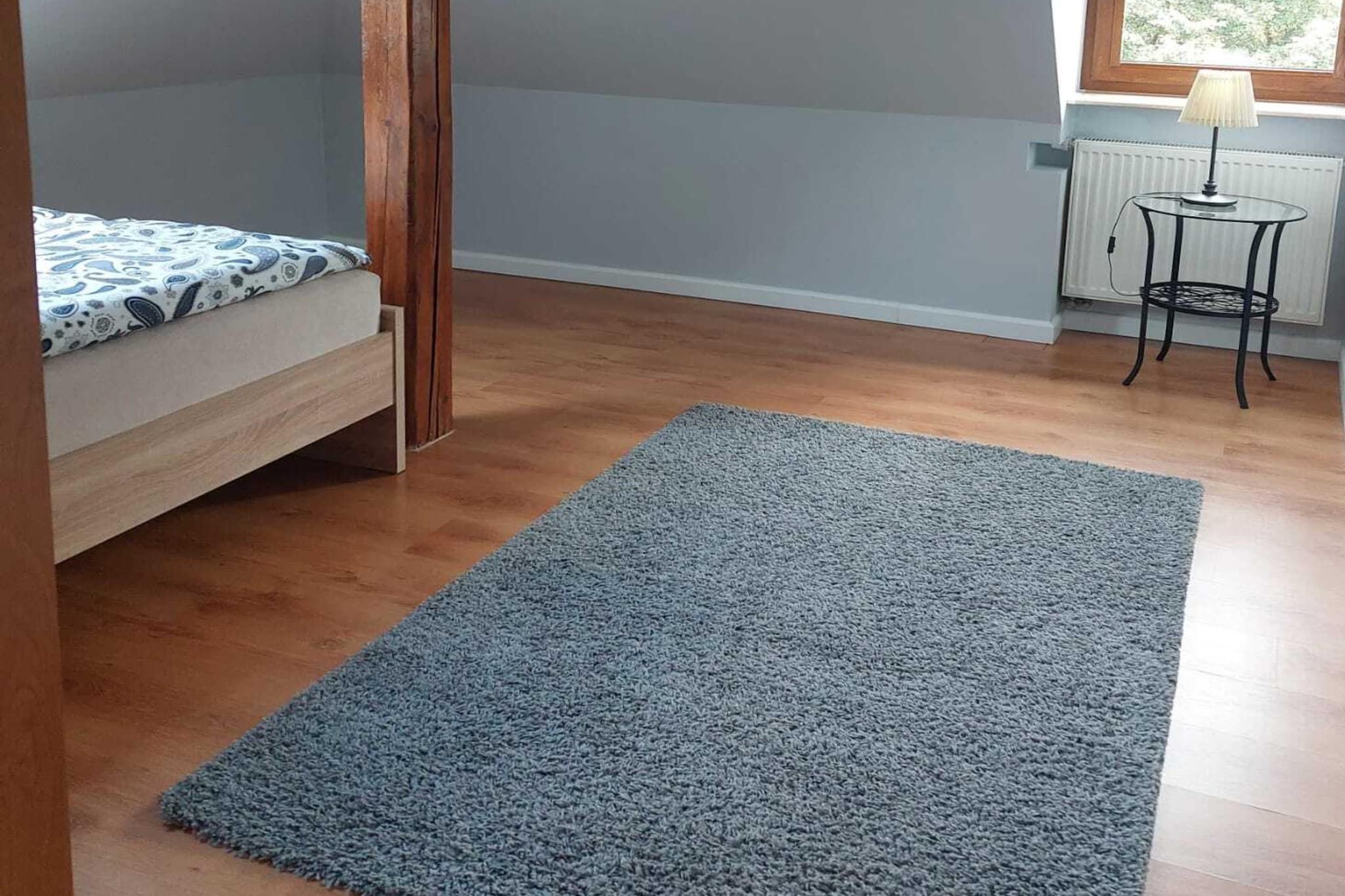 Una habitación con una cama, un tapete azul, una mesita con una lámpara y una ventana.