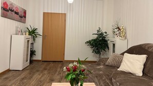 A living room with a wooden door, a brown sofa, a small table with a vase of flowers, and a white cabinet with a plant. - Apartment in Zwickau Near Horch Museum (Zwickau)
