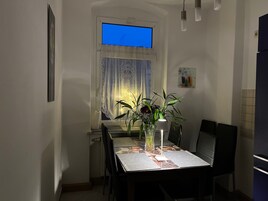 Un pequeño comedor con una mesa dispuesta para dos personas, un jarrón con flores y una ventana con cortinas.