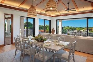 Un comedor con una mesa grande dispuesta para seis personas, rodeado de sillas blancas, y vistas al océano y la vegetación a través de grandes ventanales.