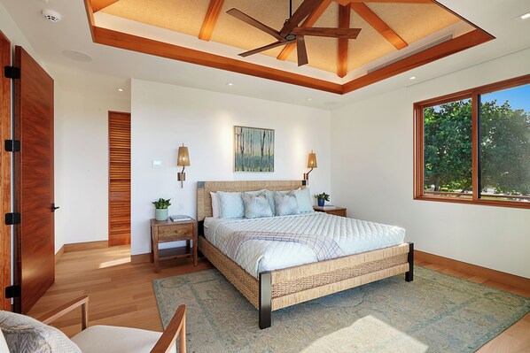 A bedroom with a large bed, wooden headboard, two bedside tables with lamps, a ceiling fan, and a window with a view of trees.