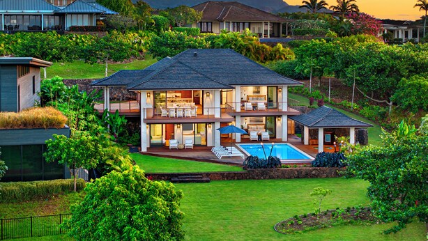 A two-story house with a large deck, a swimming pool, and lush greenery.