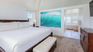 A bedroom with a large bed, a bench, a painting of a wave, a dresser, and a view of the bathroom.