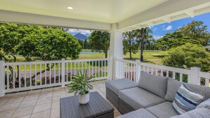 A covered outdoor area with a grey sofa and a small table, a white fence, and a view of a golf course and trees.