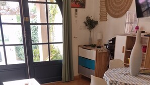 Un coin repas avec une table et des chaises, une petite table blanche sur laquelle se trouve un verre contenant une boisson rouge, un coin cuisine avec un réfrigérateur et un four à micro-ondes, et une grande fenêtre avec des rideaux.