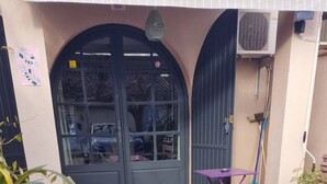 Une cour avec une chaise en plastique blanche, une table pliante violette et une nappe à rayures. On y trouve une porte noire avec une fenêtre, ainsi qu’un petit tapis vert posé sur le sol.