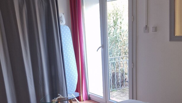 Une chambre à coucher avec un lit, une petite table, une fenêtre avec des rideaux et une vue sur la verdure à l’extérieur.