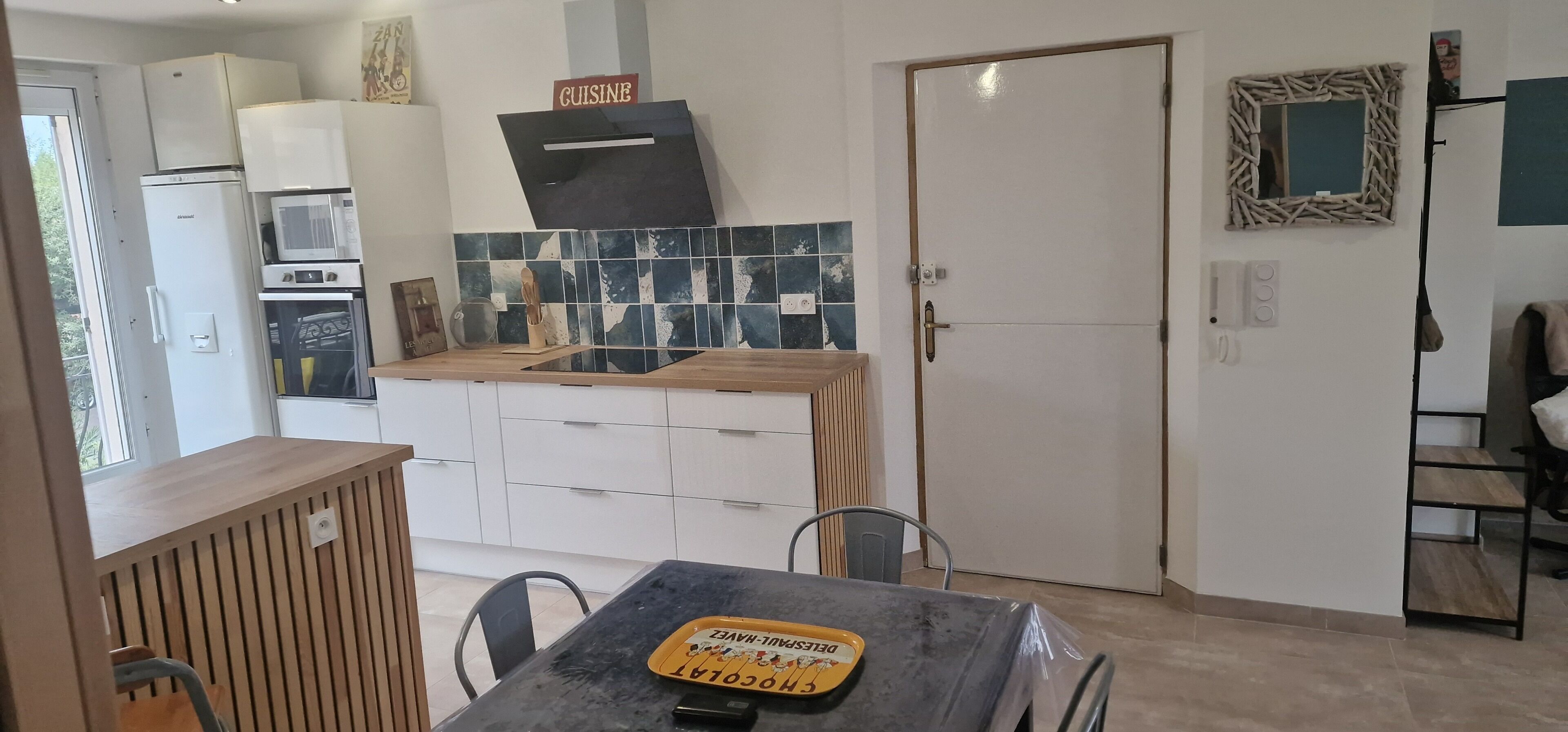 Een kleine keuken met witte kastjes, een magnetron, een zwarte afzuigkap en een betegelde spatwand.