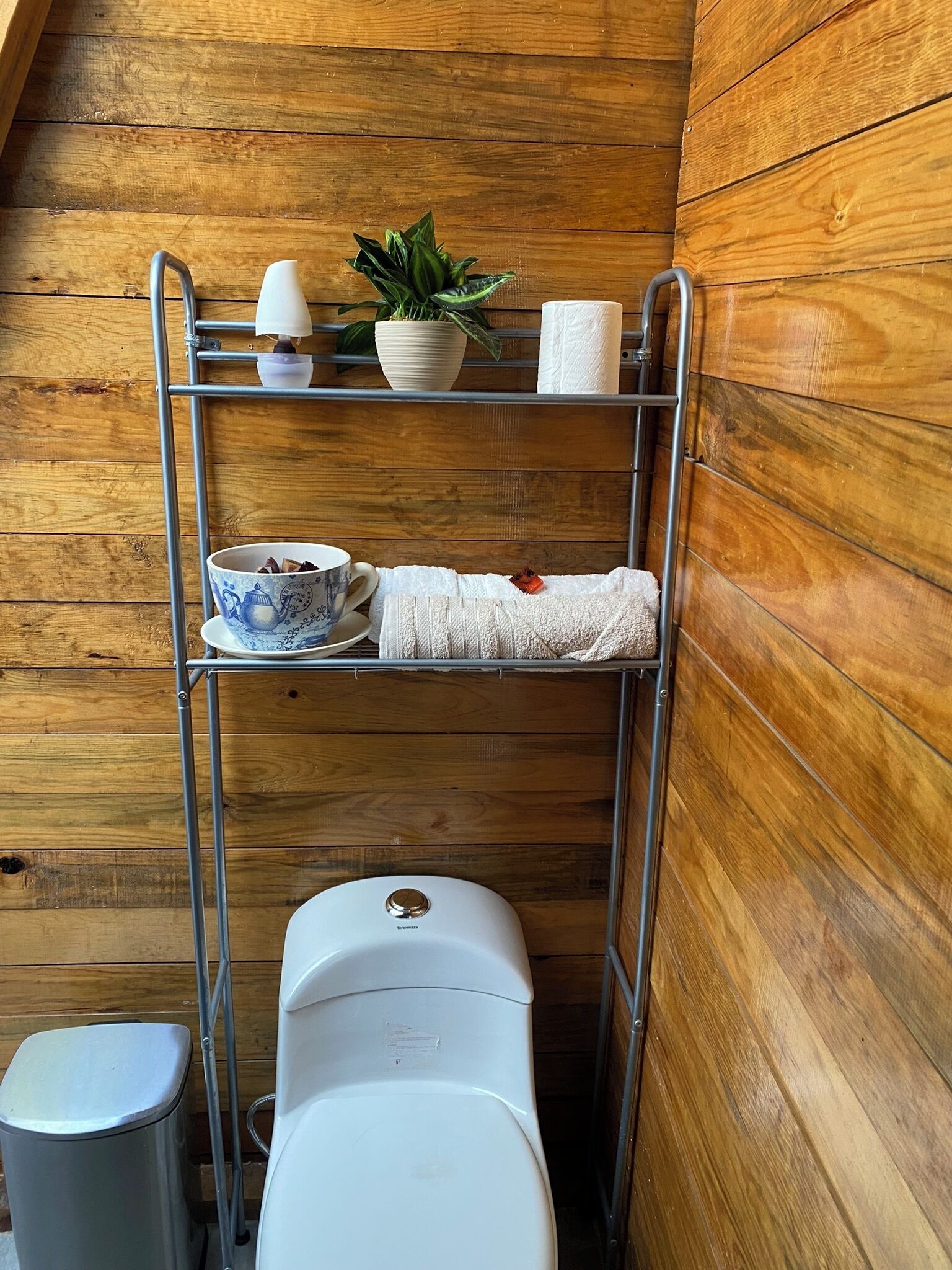 Un baño pequeño con una pared de madera, un estante metálico que sostiene un cuenco, una planta y un rollo de papel higiénico.