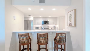 A modern kitchen with white cabinets, a stainless steel refrigerator, and a microwave. There are three wooden bar stools with patterned cushions. - 🏖️Beach Front Heated Pool 2BR Gullwing #203🏖️ (Fort Myers Beach)