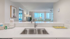 A modern kitchen with a stainless steel sink, white countertops, and a view of the living area. - 🏖️Beach Front Heated Pool 2BR Gullwing #203🏖️ (Fort Myers Beach)