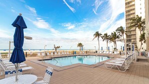 A poolside area with lounge chairs and umbrellas, a clear view of the ocean, and a high-rise building in the background. - 🏖️Beach Front Heated Pool 2BR Gullwing #203🏖️ (Fort Myers Beach)