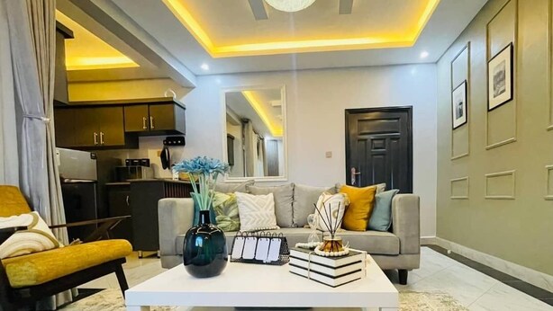 A modern living room with a sofa, coffee table, and a kitchen in the background. - Charming 1-bedroom apartment in Naalya with a serene environment. (Kampala)
