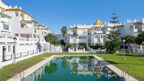 Una piscina rodeada de edificios blancos con balcones, cielo despejado y una palmera.