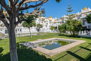 A courtyard with a swimming pool, surrounded by white buildings with yellow roofs, and a large tree in the foreground.