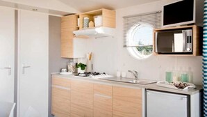 Eine kleine Küche mit Mikrowelle, Spüle und einem kleinen Kühlschrank. Ein Fenster mit Jalousien bietet Blick auf Grünfläche.