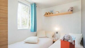 Ein Schlafzimmer mit zwei Betten, einem Fenster mit Vorhängen, einem Nachttisch mit Lampe sowie einem Regal mit Dekoration.