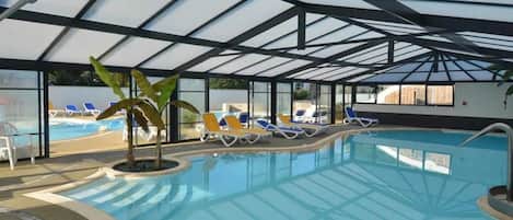 Une piscine intérieure à l’eau d’un bleu cristallin, entourée de chaises longues et dotée d’une structure de plafond en verre.