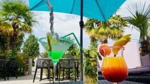 Une table avec deux boissons : un cocktail vert et une boisson rouge décorée d’un fruit, le tout placé sous un parasol bleu.