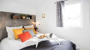 Une chambre moderne avec un lit, des oreillers, une table de chevet sur laquelle se trouve un plateau de collations, une étagère murale décorative et une fenêtre avec un rideau.