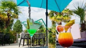 Pöydällä on kaksi juomaa: vihreä cocktail ja punainen juoma, jossa on hedelmäkoriste, ja niiden yläpuolella on sininen aurinkovarjo.