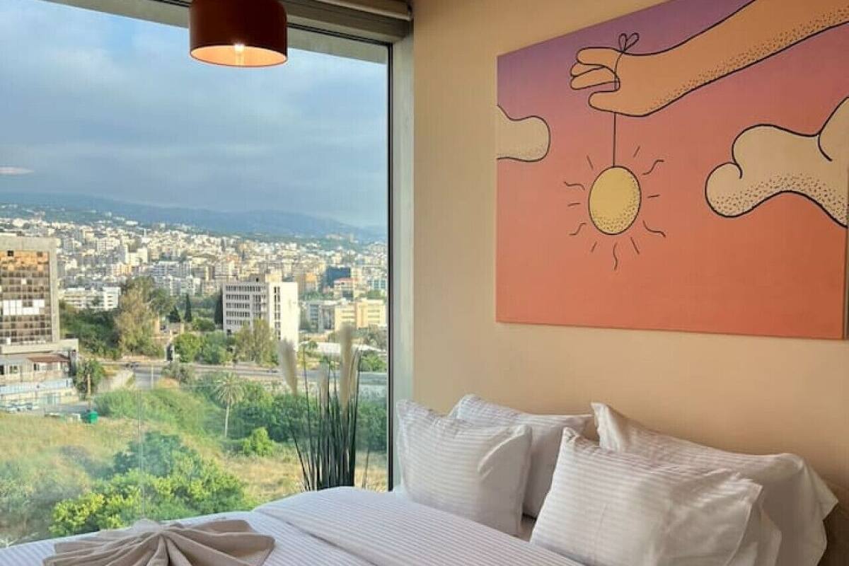 A bedroom with a large bed, a view of a cityscape, and a painting of a hand holding a sun.