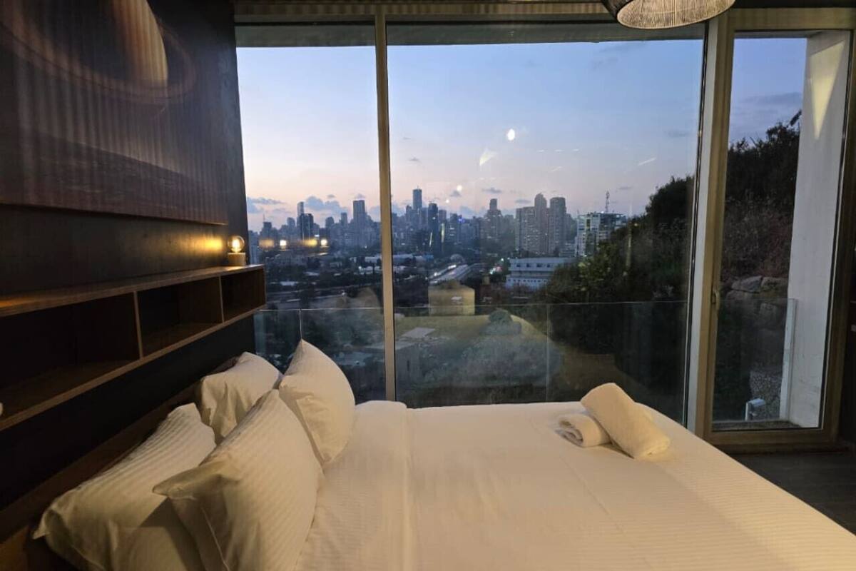 A bedroom with a large bed, a wooden headboard, and a city view through large windows.
