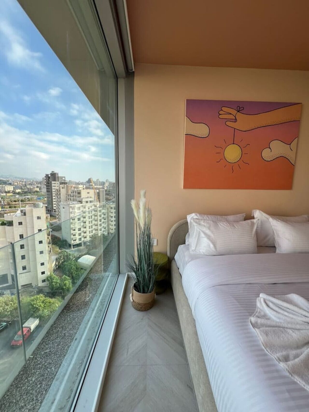 A hotel room with a large window, a bed, a painting of a hand holding a sun, and a view of the cityscape.