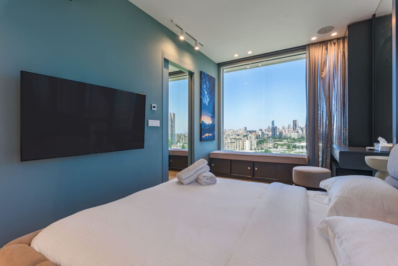 A modern hotel room with a large bed, a flat-screen TV mounted on the wall, a view of the cityscape through large windows, and a small seating area with a view.