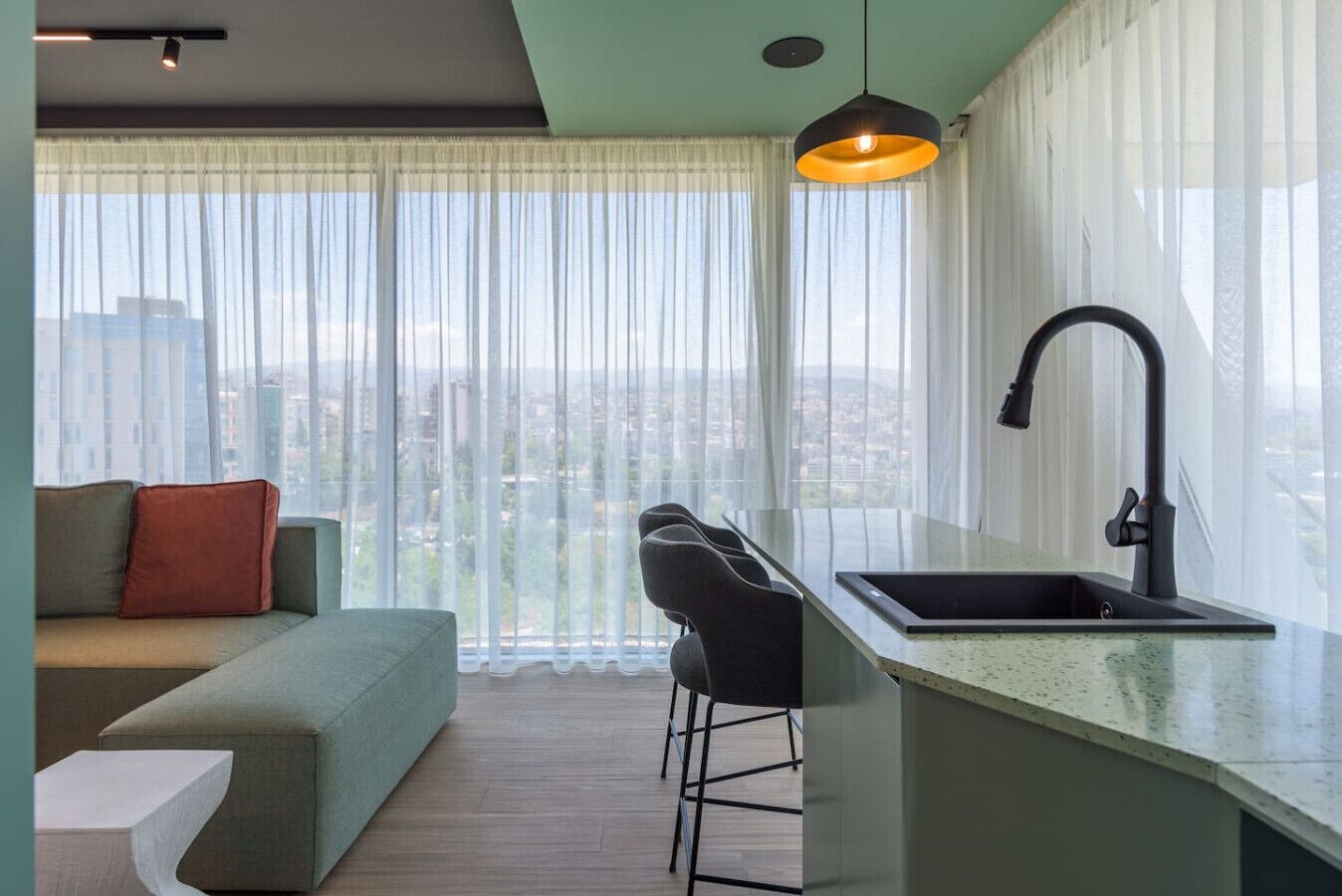 A modern kitchen with a black faucet, a bar stool, and a view of the city through sheer curtains.