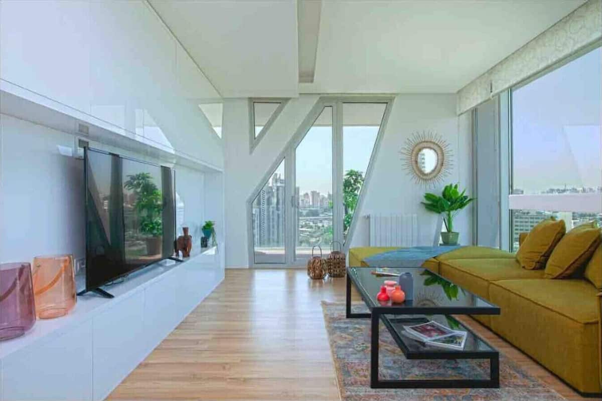 A modern living room with a sofa, coffee table, and TV, large windows with a city view, and a staircase leading to an upper floor.