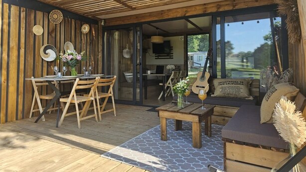 Una terraza de madera con un porche cubierto, una mesa de comedor con sillas, una mesita con un jarrón de flores y una guitarra apoyada en la pared.