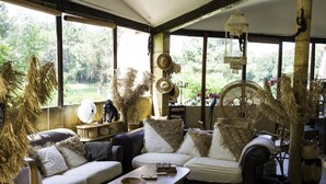 Ein gemütliches Wohnzimmer im rustikalen Stil mit einer Couch, einem Couchtisch und Korbmöbeln. Große Fenster bieten Blick auf viel Grün draußen.