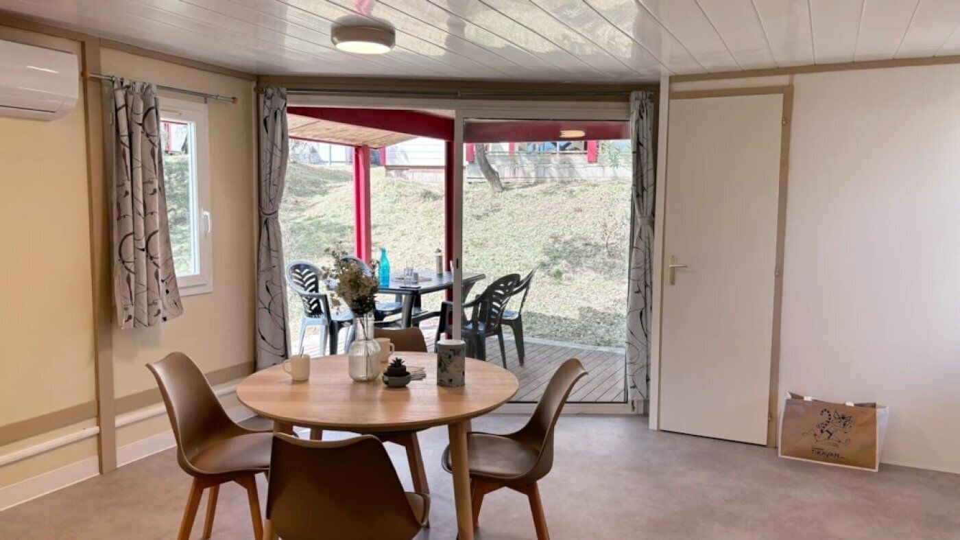 Une pièce avec une table ronde en bois et quatre chaises, une porte-fenêtre coulissante donnant sur l’extérieur, et une petite table sur laquelle se trouvent un vase et une plante.