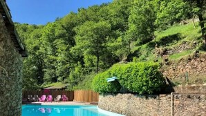 A swimming pool with stone walls, surrounded by greenery and a wooden deck with chairs. - ZEcamping piscine chiens acceptés jacuzzi locatif BBQ 30m2 4 pers (Saint-Constant-Fournoulès)