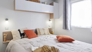 A bedroom with a bed, pillows, a nightstand, a wall-mounted shelf, and a window with curtains.