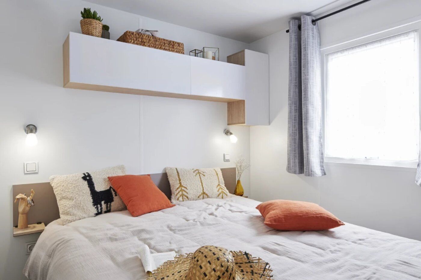 A bedroom with a bed, pillows, a nightstand, a wall-mounted shelf, and a window with curtains.