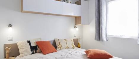 A bedroom with a bed, pillows, a nightstand, a wall-mounted shelf, and a window with curtains.