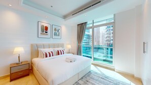 A modern hotel room with a large bed, a nightstand, a window with a city view, and two framed artworks on the wall.