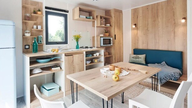 A modern kitchen with wooden cabinetry, a dining table with chairs, and a blue sofa in a living area. - ZEcamping 4 étoiles animé 500m plage piscine couverte mobil-home 25m2 (Morsalines)