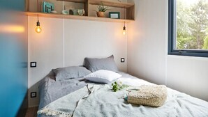 A bedroom with a bed, a window, and a shelf with decorative items. - ZEcamping 4 étoiles animé 500m plage piscine couverte mobil-home 25m2 (Morsalines)