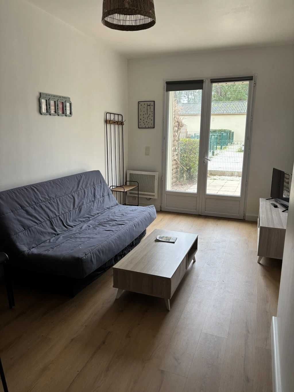 A modern living room with a grey sofa, a wooden coffee table, and a TV stand. There is a large window with a view of a garden and a building outside.