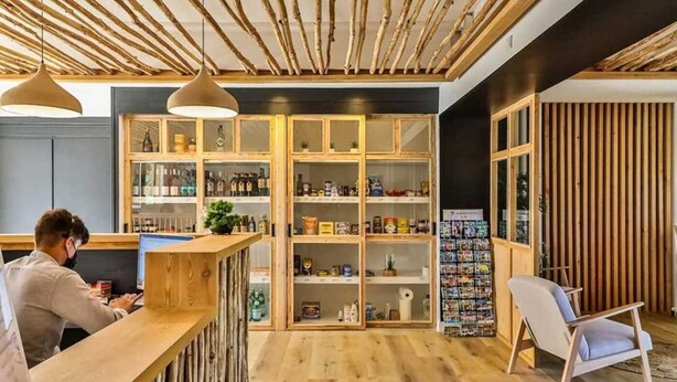 Un espace bar moderne avec des étagères en bois remplies de divers articles, un coin salon avec un fauteuil, et une personne qui travaille au bar.