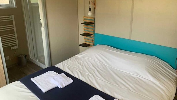 A hotel room with a bed, a towel, a shelf with books, and a light fixture.