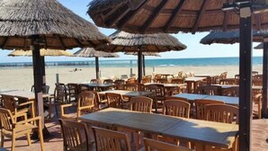 Un área de estar al aire libre con mesas y sillas de madera, sombrillas de paja y vista a la playa.