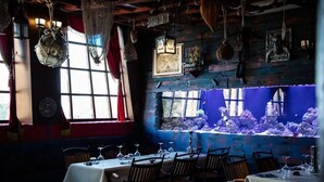 A restaurant with a rustic interior, featuring wooden walls, hanging lanterns, and a large aquarium window. Tables are set for dining.