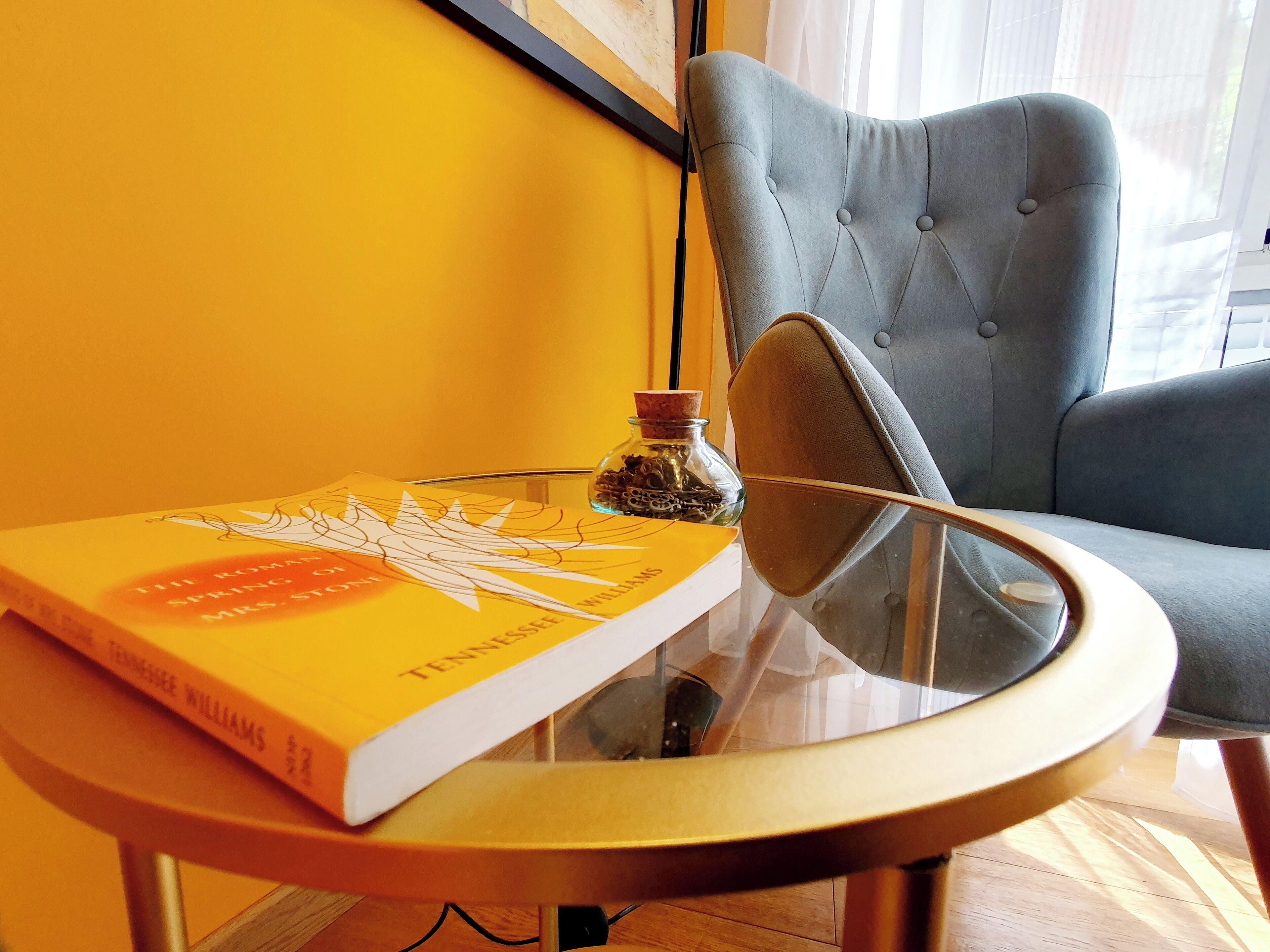 Un livre intitulé « Le printemps romain, Madame Stoddard » est posé sur une table en verre, à côté d’un petit pot d’épices et d’un fauteuil recouvert d’un tissu gris.