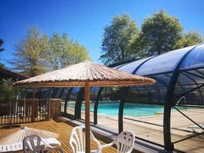 Área da piscina com guarda-sol de palha, cadeiras de plástico branco e estrutura transparente para a piscina.
