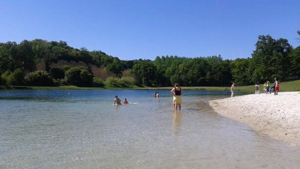 A beach with clear water, people swimming and standing on the shore, and a forested hill in the background.
