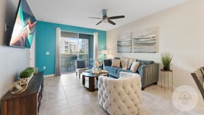 A modern living room with a teal accent wall, a ceiling fan, a large flat-screen TV, a sofa set, a tufted ottoman, and a small side table with a plant. - 3120-105 Resort Pool View Disney Universal Orlando (Kissimmee)